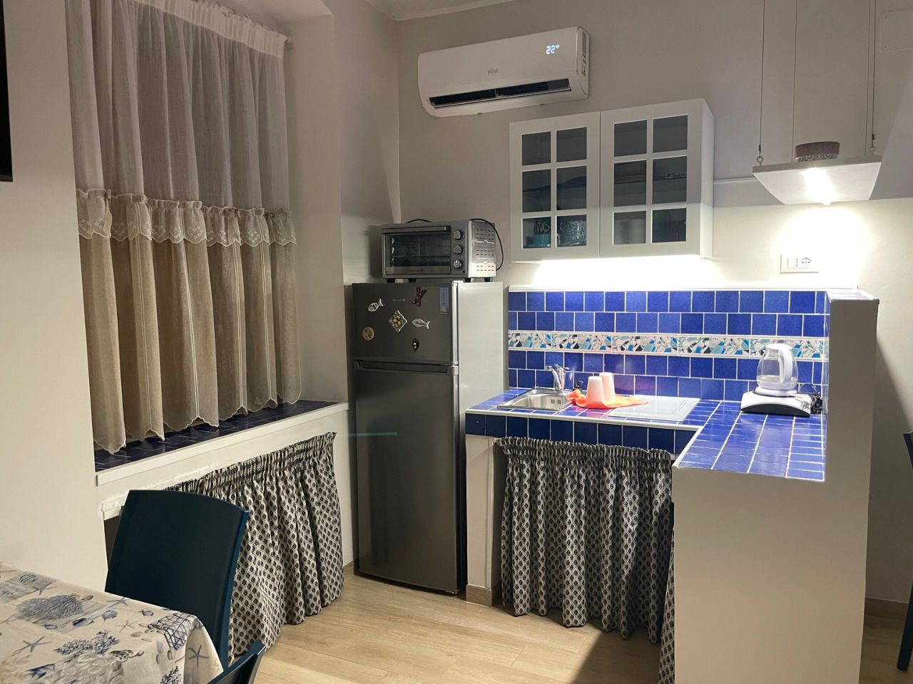 Angolo cottura attrezzato con piastrelle blu, frigorifero, forno e condizionatore d'aria - Casa Vacanze BluMare Taranto